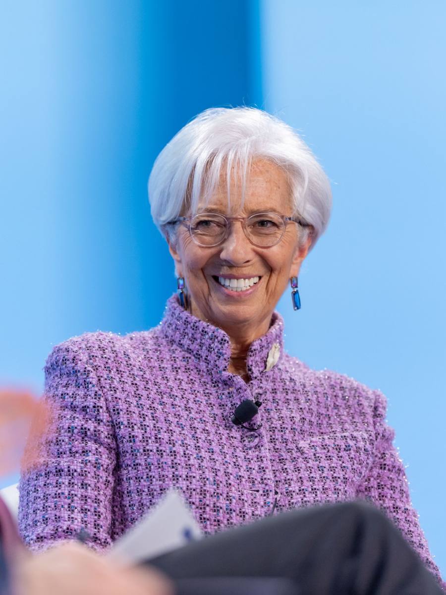 Christine Lagarde, zâmbind, cu ochelari și sacou mov, în fața unui fundal albastru